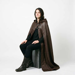 Woman seated on studio block wearing flowing brown leather medieval mantle displaying loose drapey hem and cowl hood