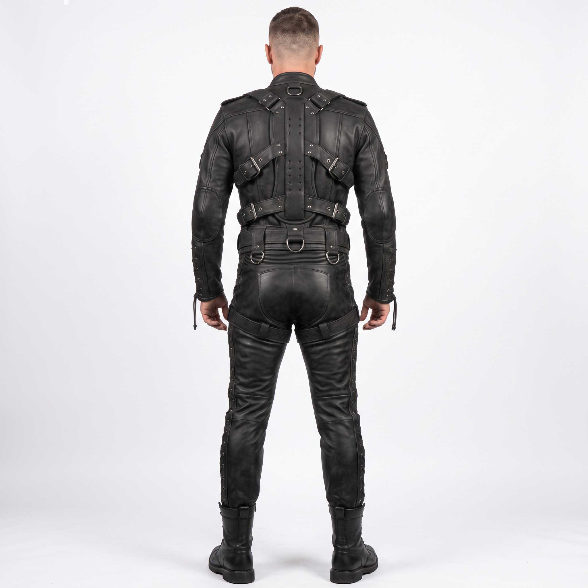 Back view of man modeling black leather bondage suit showing articulated spine harness, metal D rings, rear cross straps, and lace up legs