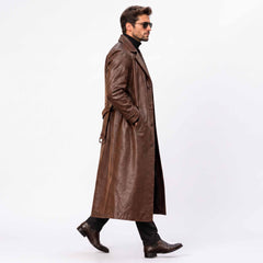 Side profile walking shot of man wearing tailored long brown leather trench coat with hands in pockets against white background