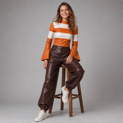 Woman seated on a high wooden stool showcasing the gathered leather folds and tailored waist construction of the brown leather joggers and striped top